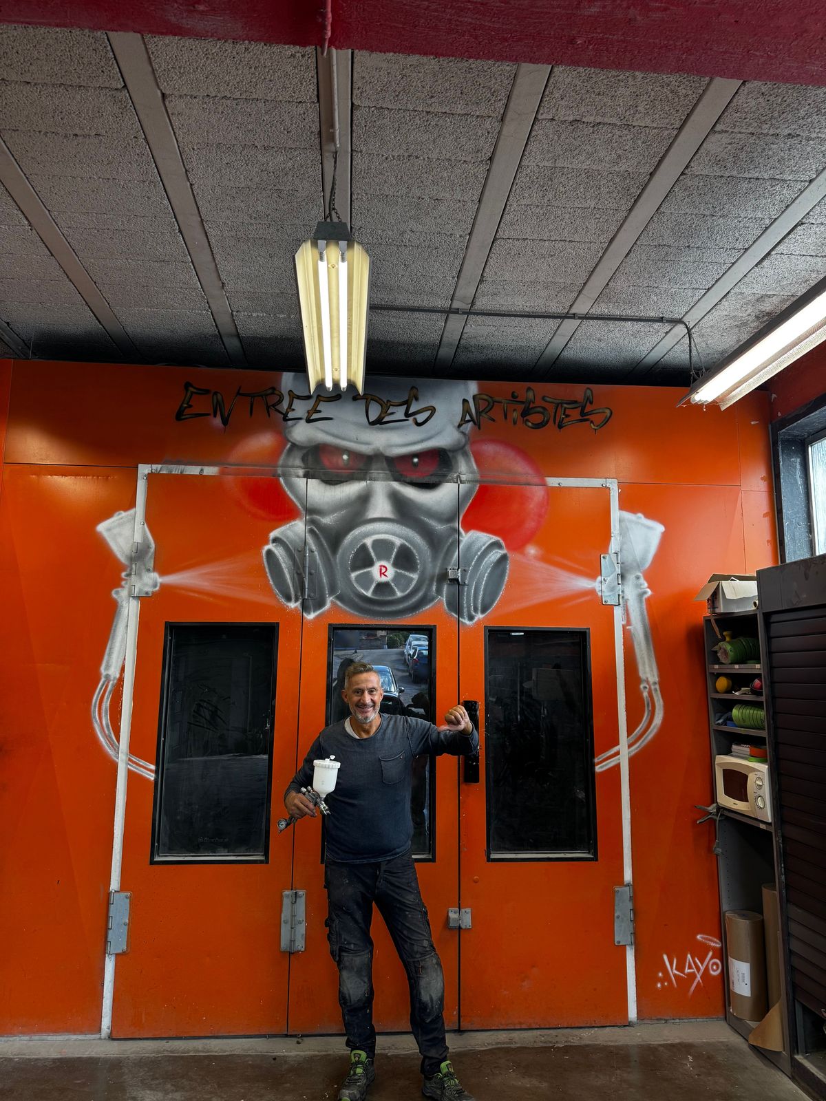 Edouardo Ricciolini devant la cabine de peinture « Entrée des artistes », décorée d'une fresque de masque à gaz, un pistolet à peinture à la main.